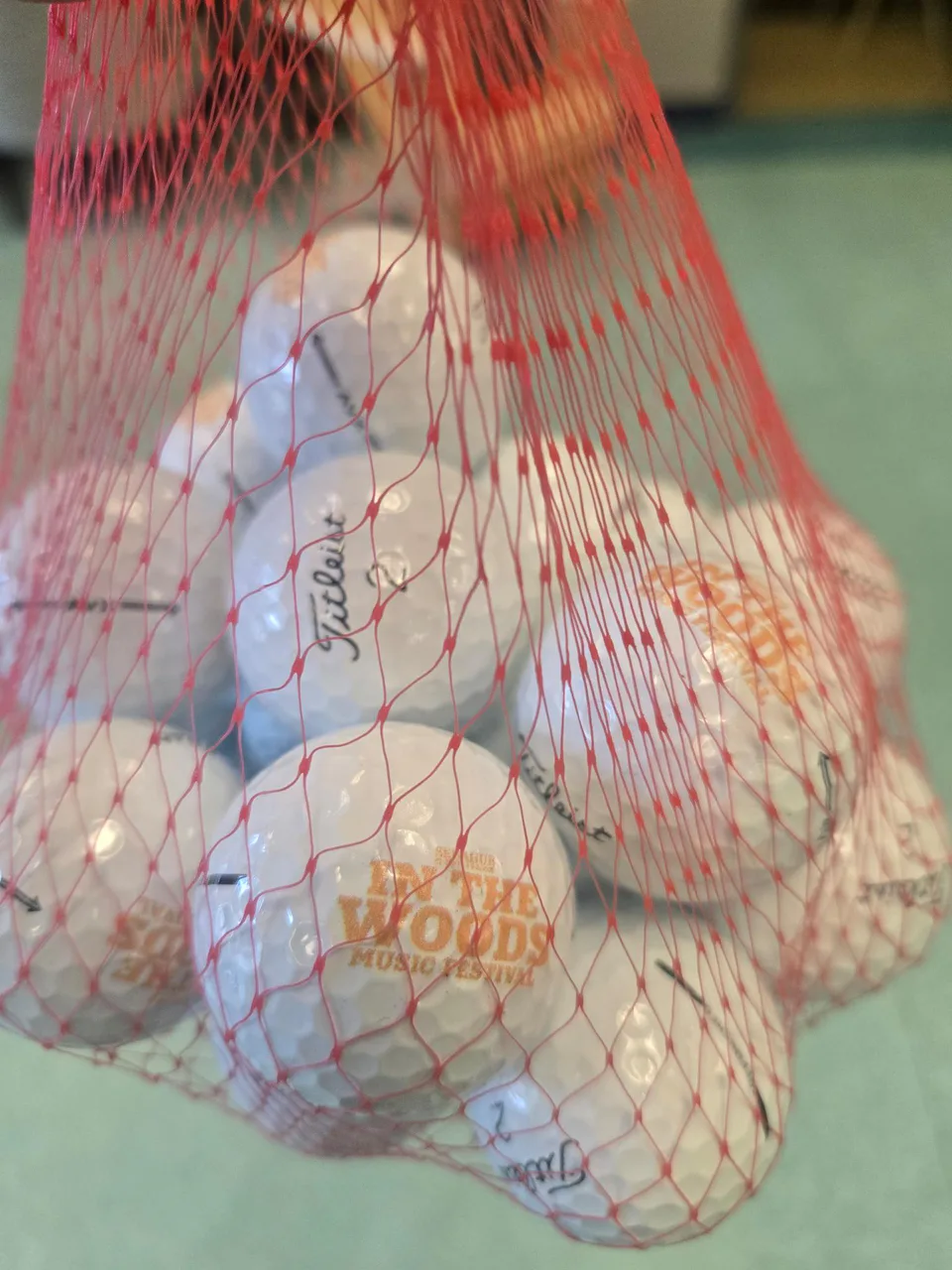 A red mesh bag holding many white golf balls, including balls carrying the orange In the Woods logo